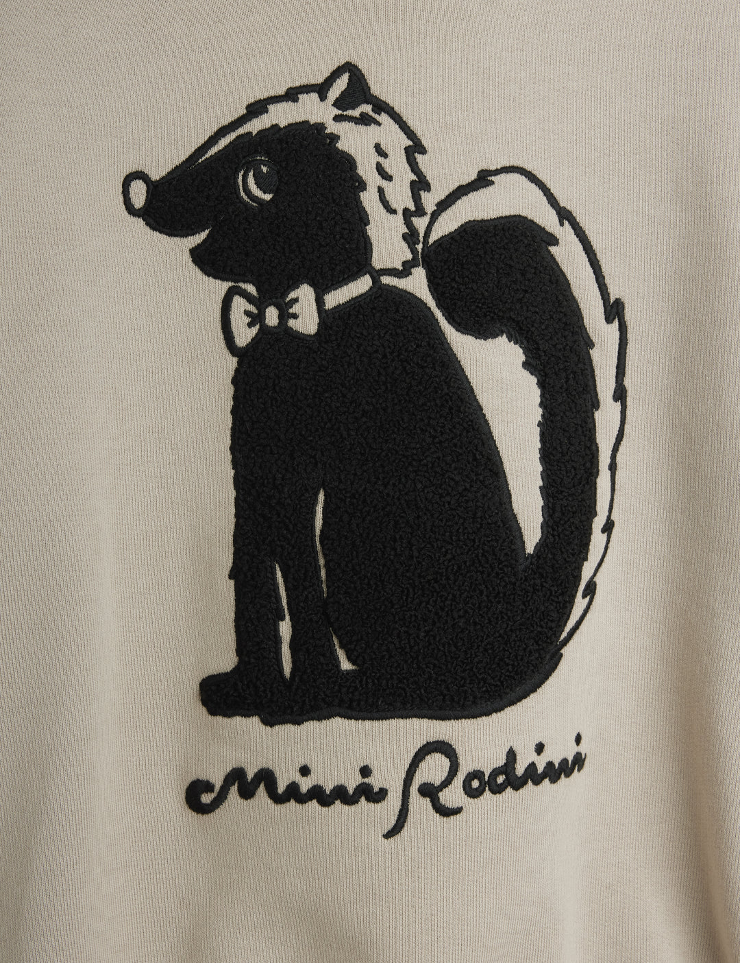 Close-up of a cream-colored garment featuring a black embroidered skunk graphic wearing a bow tie, with the text 'Mini Rodini' embroidered below.