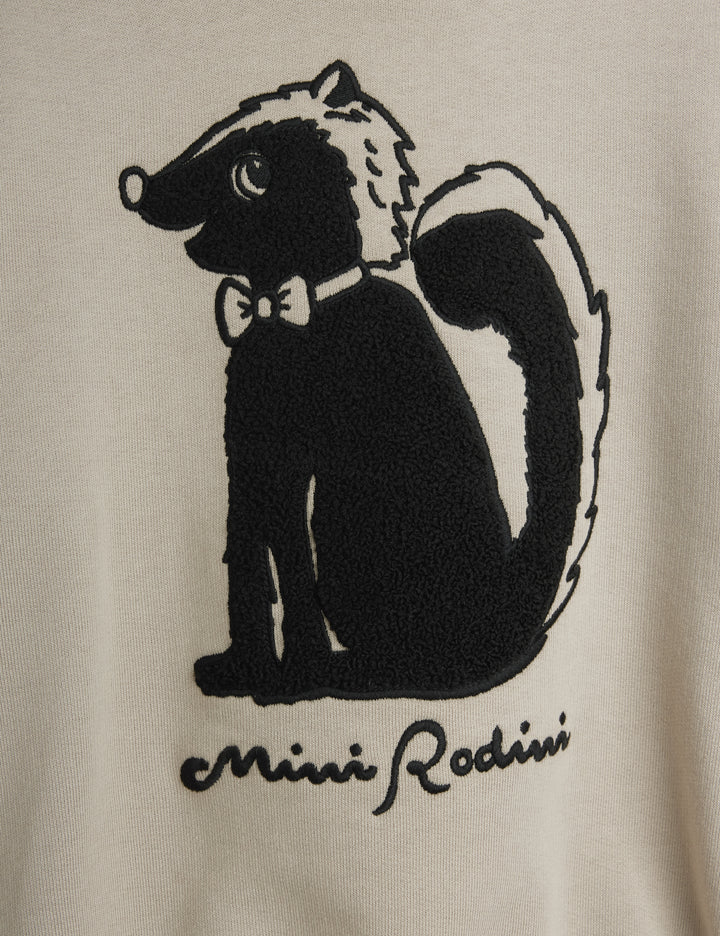 Close-up of a cream-colored garment featuring a black embroidered skunk graphic wearing a bow tie, with the text 'Mini Rodini' embroidered below.