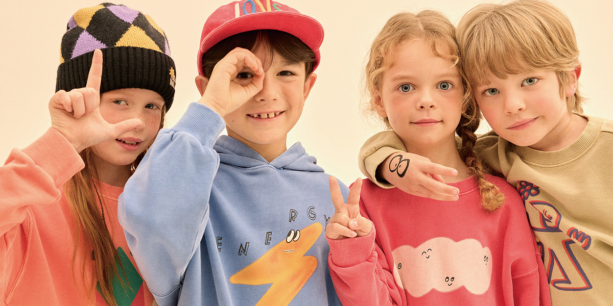 Four children wearing colorful casual clothing and hats, posing together against a plain beige background. The children are dressed in pastel and bright colors, including a light blue hoodie, a pink sweatshirt, a red cap, a peach sweatshirt, and a black-and-purple patterned beanie.