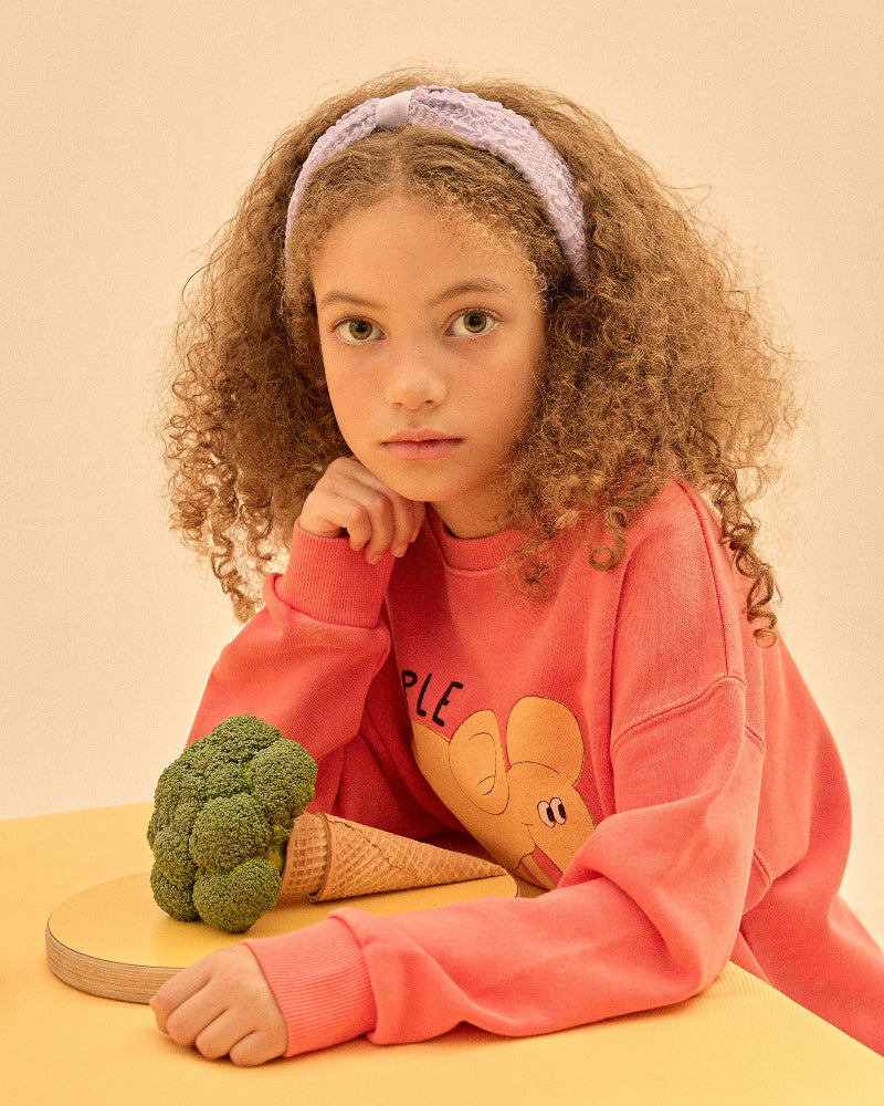 Child wearing a pink sweatshirt with a heart design, sitting next to a broccoli and ice cream cone on a yellow surface.