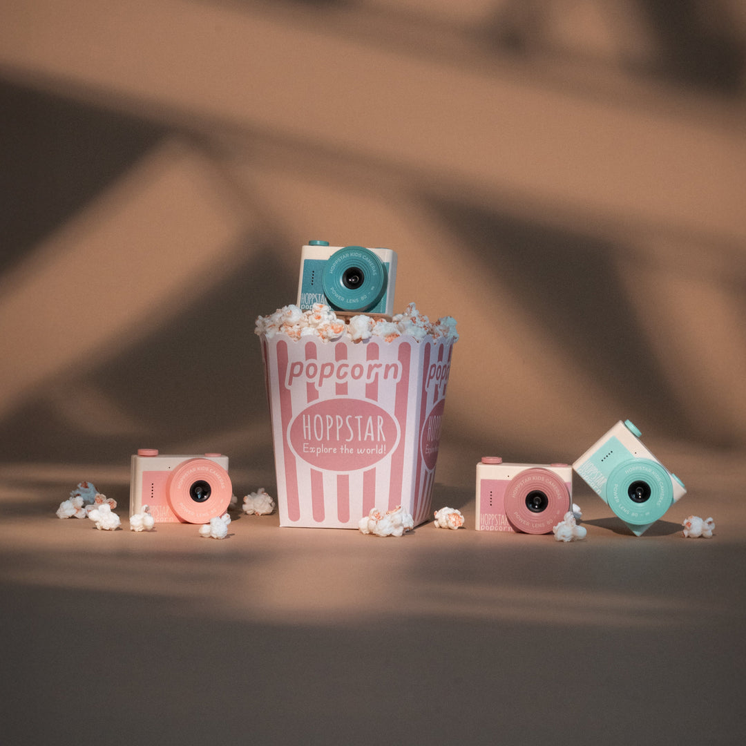 A set of miniature retro-style film cameras in pink, blue, and white, styled with a small pink-and-white striped popcorn bucket and scattered popcorn kernels on a dark surface with warm, dramatic shadows.